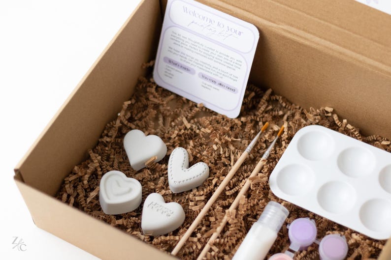 May include: A cardboard box filled with craft supplies for painting ceramic hearts. The box contains four white ceramic hearts, two paintbrushes, a small container of white paint, a small container of purple paint, and a white paint palette with six wells.