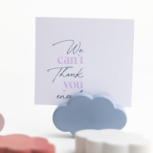 May include: A set of three wooden cloud-shaped note holders in pink, red, and blue. The blue cloud holder has a white card with the text "We can't thank you enough" written in black and purple.