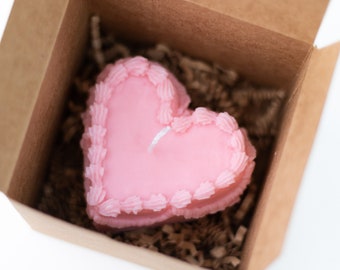 May include: A pink heart-shaped candle with a white wick sits in a brown cardboard box with brown paper shreds.