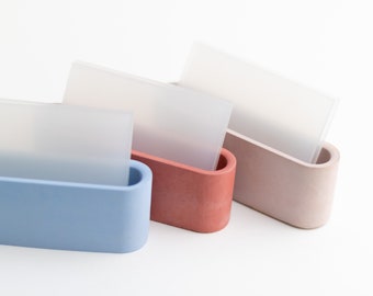 Könnte beinhalten: Drei moderne, minimalistische Visitenkartenhalter in Blau, Rot und Beige. Jeder Halter besteht aus einem glatten, strukturierten Material und hat eine abgerundete, ovale Form.