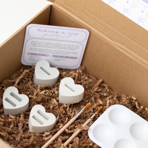May include: A cardboard box filled with craft supplies for painting. The box contains four white heart-shaped ceramic pieces, two paintbrushes, a white paint palette with six wells, and brown packing paper.