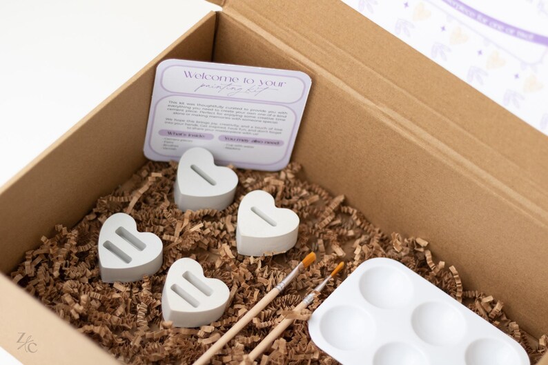 May include: A cardboard box filled with craft supplies for painting. The box contains four white heart-shaped ceramic pieces, two paintbrushes, a white paint palette with six wells, and brown packing paper.