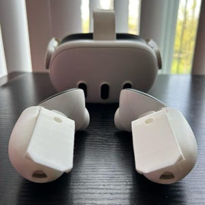 Könnte beinhalten: Ein weißes Virtual-Reality-Headset mit einem zentralen Kopfband und drei schwarzen Sensoranschlüssen an der Vorderseite, begleitet von zwei weißen Handcontrollern mit flachen, rechteckigen weißen Aufsätzen an der Oberseite.