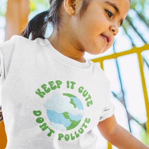 Może przedstawiać: Biała koszulka z zielonym napisem "KEEP IT CUTE DON'T POLLUTE". Projekt zawiera globus z niebieskimi oceanami i zielonymi kontynentami. Koszulka jest noszona przez małe dziecko.