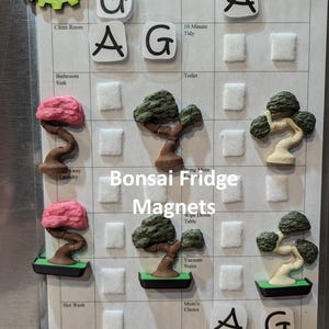 May include: A set of colorful magnetic bonsai tree fridge magnets. The magnets are arranged on a white grid with labels for various household chores. The labels include "Clean Room", "Bathroom Sink", "10 Minute Tidy", "Toilet", "Away Laundry", "Bring Mom", "Wipe Down Table", "Vacuum Stairs", "Hot Wash", "Mom's Choice", "Weeding", and "Water Plants".