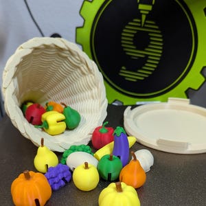 May include: A white wicker basket overflowing with colourful 3D printed fruits and vegetables, including a red apple, yellow bell pepper, green pear, orange pumpkin, purple grapes, yellow lemon, green apple, orange pear, red pepper, yellow banana, purple aubergine, and green broccoli.