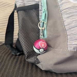 May include: A pink and white Pokeball keychain with a pink heart on the front, attached to a silver key ring. The keychain is hanging from a zipper on a gray and white backpack.