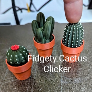 May include: Three small potted cactus plants, one with a red flower on top, are shown on a grey surface. The text "Fidgety Cactus Clicker" is visible below the plants.