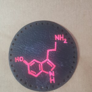 Puede incluir: Un parche de cuero negro con una estructura química rosa de serotonina, un neurotransmisor. La estructura química está delineada en rosa e incluye las letras "NH2", "HO" y "NH".