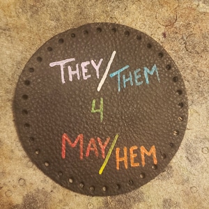 May include: Black leather circle with white, blue, yellow, and orange writing. The writing says "They/Them 4 May/Them".