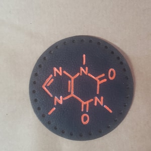 Puede incluir: Un posavasos de cuero negro con una estructura química naranja de cafeína impresa en él. El posavasos tiene un círculo de pequeños agujeros alrededor del borde.