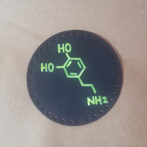 May include: A black round patch with a white stitched border. The patch has a green chemical structure with the text "HO", "HO", and "NH2" written on it.