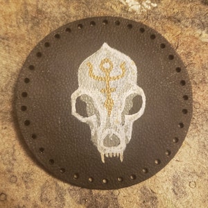 May include: A black leather coaster with a white and gold painted animal skull with a symbol on its forehead.