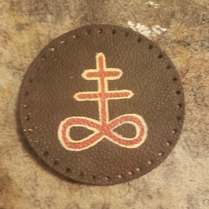 May include: A round, dark brown leather patch with a red and white symbol in the center. The symbol is a cross above an infinity symbol. The edge of the patch has evenly spaced holes.