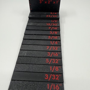 May include: Black stepped gauge with red measurements. The top block is 3" x 2" x 1". The gauge has measurements from 1/16" to 3/4". The tool is used for measuring and checking dimensions.