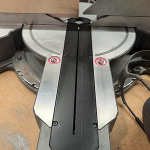 May include: A miter saw with a silver blade guard and a black base. The saw is designed for cutting wood, with a wooden board positioned on the saw's surface. Safety symbols are visible.