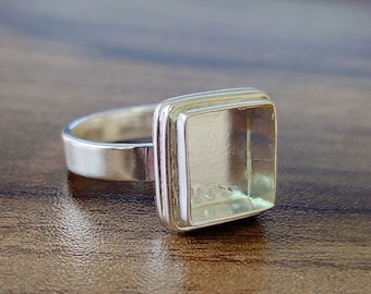 May include: A silver ring with a square-shaped clear stone setting. The ring is on a wooden surface.