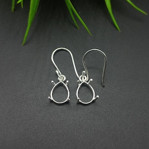 May include: A pair of silver teardrop-shaped earring findings. Each earring has a wire hook and a decorative setting with small prongs around the teardrop shape. The earrings are displayed against a dark gray background.