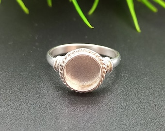 Könnte beinhalten: Ein silberner Ring mit einer runden Zargenfassung. Die Zarge ist glatt und hat einen leicht erhöhten Rand. Der Ringarm ist dünn und hat ein seilartiges Design.