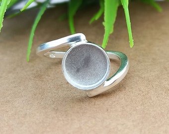 Könnte beinhalten: Ein silberner Ring mit einer runden Fassung für einen Cabochon-Stein. Der Ring ist verstellbar und hat ein schlichtes, modernes Design.