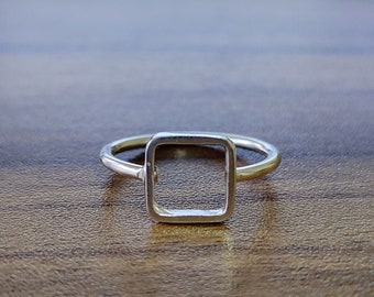 May include: A silver ring with a square-shaped open design. The ring is on a wooden surface.