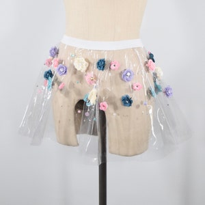 May include: A sheer, white tulle skirt with a variety of colorful fabric flowers attached. The skirt is on a mannequin and has a white waistband.