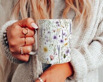 Può includere: Una tazza in ceramica bianca con un motivo floreale con fiori selvatici viola, rosa e gialli. La tazza è tenuta da una mano.