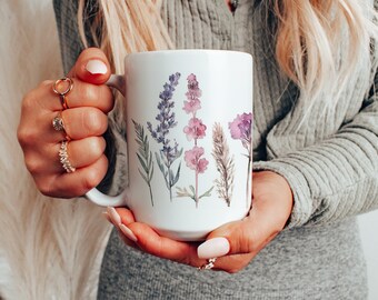 Può includere: Una tazza in ceramica bianca con un design ad acquerello di fiori viola e rosa. La tazza ha un manico ed è tenuta dalle mani di una persona.