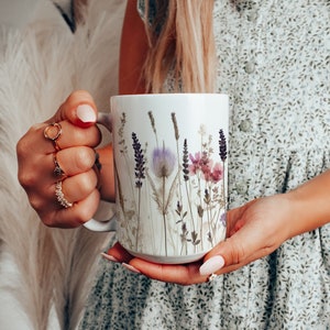 Könnte beinhalten: Weiße Keramiktasse mit einem gepressten Blumendesign mit violetten, rosa und grünen Blumen. Die Tasse hat einen Henkel und wird von einer Hand gehalten.