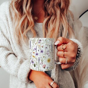 Peut inclure: Une tasse en céramique blanche avec un motif floral composé de fleurs violettes, roses et jaunes. La tasse est tenue par une main.