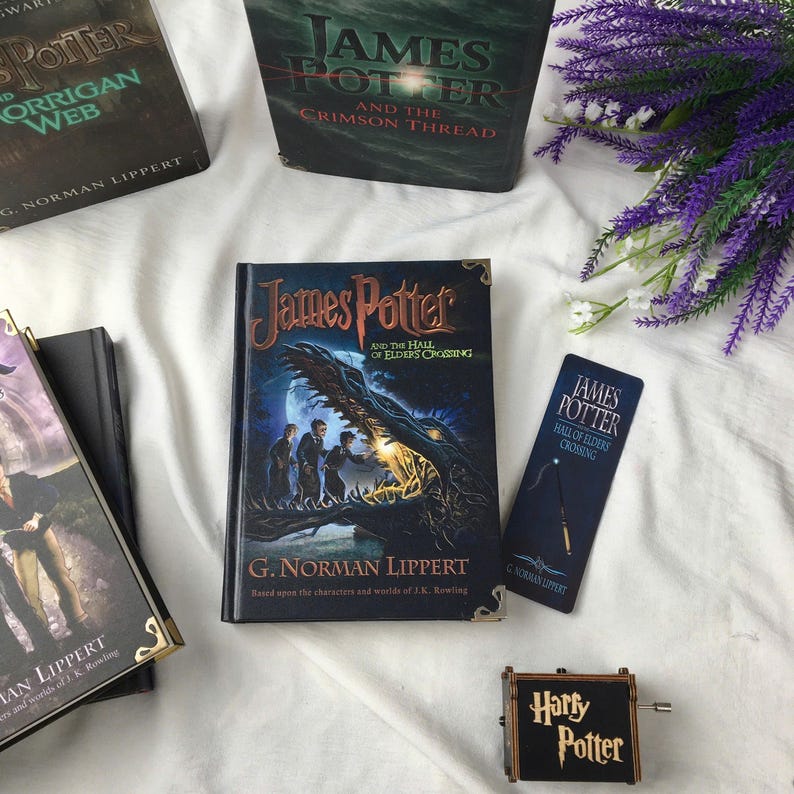 May include: A collection of "James Potter" books, a bookmark, and a music box are arranged on a white cloth. The books feature dark covers with illustrations and text. A small music box with "Harry Potter" is also present, along with purple flowers.