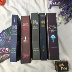 May include: A selection of hardcover books, including titles such as "James Potter and the Hall of Elders Crossing" and "James Potter and the Morrigan Web." The books are in shades of blue, burgundy, and green. A small "Harry Potter" music box is also visible.