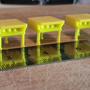 May include: Three yellow plastic miniature tables with four legs each. The tables are arranged in a row on a wooden surface with a ruler underneath.