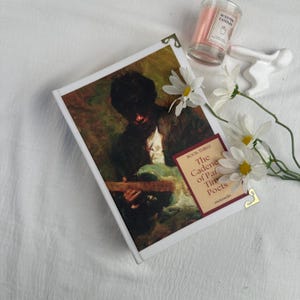 Pode incluir: Um livro de capa dura intitulado "The Cadence of Parisian Poets" com uma pintura vintage na capa. O livro &eacute; adornado com protetores de canto dourados e repousa sobre uma superf&iacute;cie branca, acompanhado por margaridas e uma vela perfumada.