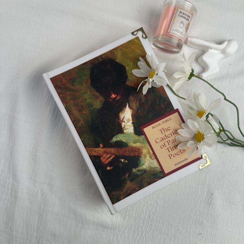 Pode incluir: Um livro de capa dura intitulado "The Cadence of Parisian Poets" com uma pintura vintage na capa. O livro &eacute; adornado com protetores de canto dourados e repousa sobre uma superf&iacute;cie branca, acompanhado por margaridas e uma vela perfumada.