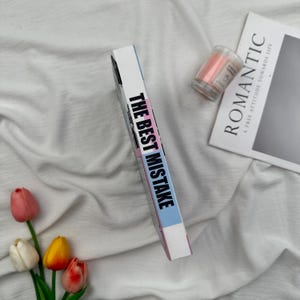 Puede incluir: Un libro titulado "THE BEST MISTAKE" con una portada rosa, azul y blanca, descansa sobre una superficie de tela blanca. Tambi&eacute;n hay una peque&ntilde;a vela rosa en un tarro de cristal y una revista con la palabra "ROMANTIC". Un ramo de tulipanes a&ntilde;ade color.