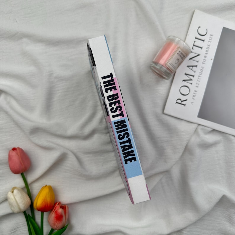 Puede incluir: Un libro titulado "THE BEST MISTAKE" con una portada rosa, azul y blanca, descansa sobre una superficie de tela blanca. Tambi&eacute;n hay una peque&ntilde;a vela rosa en un tarro de cristal y una revista con la palabra "ROMANTIC". Un ramo de tulipanes a&ntilde;ade color.