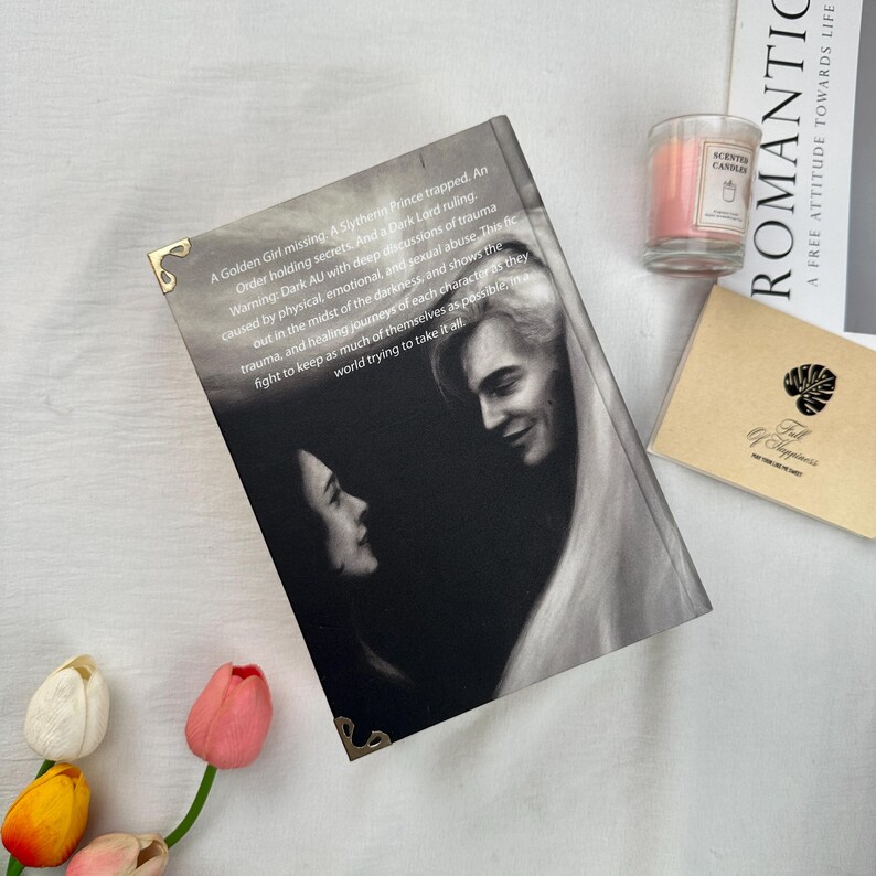 May include: A book with a black and white image of two people on the cover. The book has gold corner protectors. Text on the cover describes the story as a dark AU with discussions of trauma. A scented candle and a small card are also in the image.