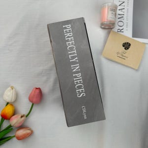 May include: A gray rectangular box with the text "PERFECTLY IN PIECES" in white. The box is next to a small candle in a glass jar, a small card, and a book. Several colorful tulips are in the lower left corner.
