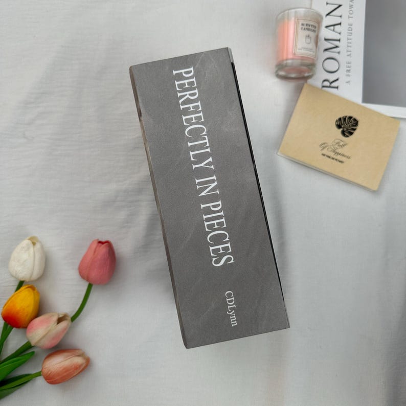 May include: A gray rectangular box with the text "PERFECTLY IN PIECES" in white. The box is next to a small candle in a glass jar, a small card, and a book. Several colorful tulips are in the lower left corner.