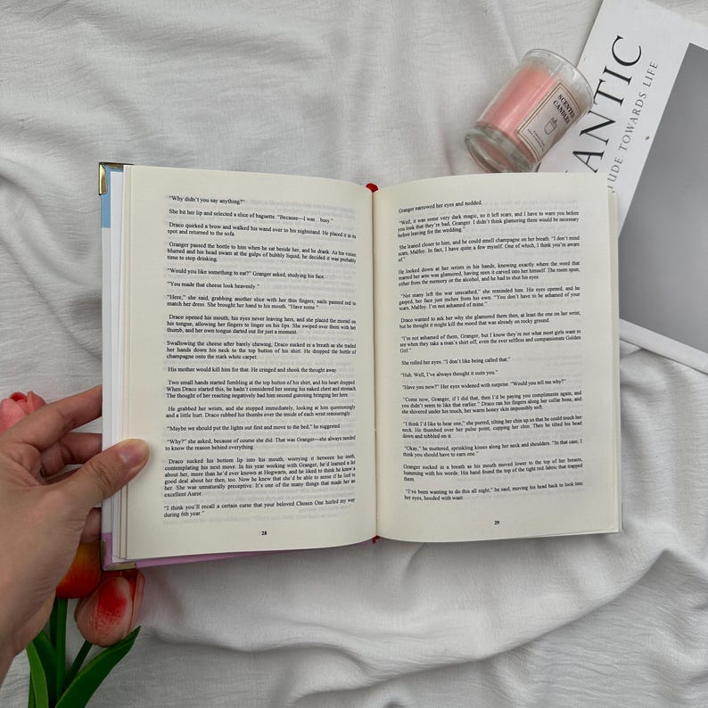 Puede incluir: Un libro abierto con texto en p&aacute;ginas color crema, sostenido por una mano. Una vela rosa en un tarro de cristal y una revista con la palabra "ANTIC" est&aacute;n en el fondo. El libro tiene una cinta roja como marcador.