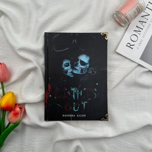 May include: A hardcover book with a dark cover featuring two skulls and the title "Lights Out" by Navessa Allen. The book has gold corner protectors and is set on a white fabric with a candle and a magazine.
