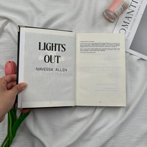 May include: An open book with the title "LIGHTS OUT" by Navessa Allen. The book has a black cover with gold corners. A pink candle and a magazine are in the background.