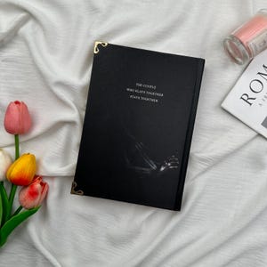 May include: A black journal with gold corner protectors and the text "THE COUPLE WHO SLAYS TOGETHER STAYS TOGETHER" in white. The journal is on a white, textured surface with a candle and a magazine in the background.