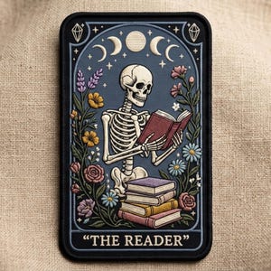 May include: Embroidered patch featuring a skeleton reading a book, surrounded by flowers and moons. The design includes the text "THE READER" at the bottom, set against a dark blue background with a black border. The patch is rectangular.