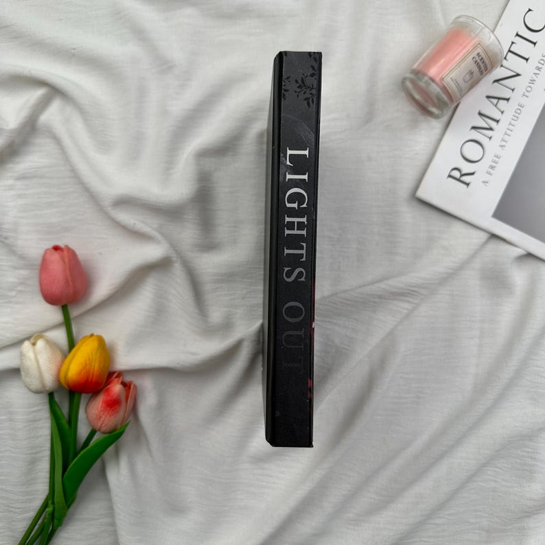 May include: A black book titled "LIGHTS OUT" with white lettering. The book is positioned vertically, with a floral design on the spine. A small pink candle in a glass jar and a magazine with the word "ROMANTIC" are also visible.