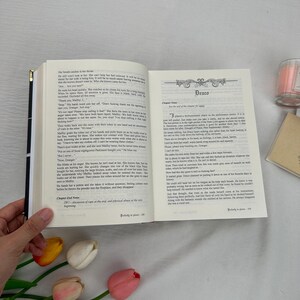 May include: An open book with text on the pages, held by a hand. The book's title page reads "Draco." A candle and a small notepad are in the background. The book is open to a chapter with text.