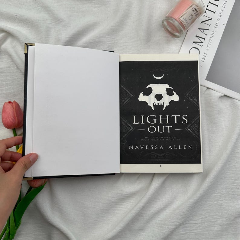 May include: An open book with a black cover and gold-colored corner accents. The right page displays the cover of "LIGHTS OUT" by Navessa Allen, featuring a skull design. A pink candle and a magazine are in the background.