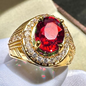 May include: A gold-toned ring featuring a large, oval-cut, deep red gemstone as the centerpiece. The gemstone is surrounded by a halo of smaller, clear stones. The band has an intricate, textured design.