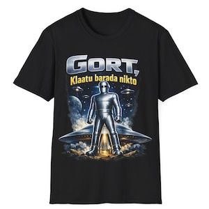 Może przedstawiać: Czarny t-shirt z krótkim rękawem z nadrukiem graficznym przedstawiającym metalicznego robota stojącego przed latającym spodkiem. Nad robotem znajduje się napis GORT, Klaatu barada nikto, a w tle małe latające spodki i planeta.